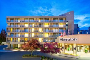 Photo of Sheraton Vancouver Airport Hotel. POV: facing entrance of hotel. Dark blue sky, lights on in hotel. Out  front, two trees with dark red leaves.
