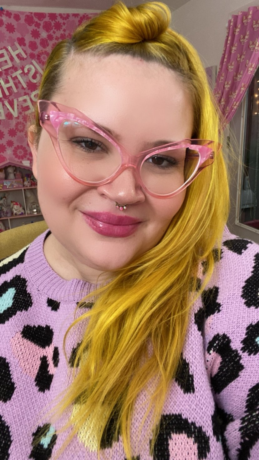 Caucasian person with pink rimmed glasses, high pony tail, blond hair, and purple sweater with leopard print pattern