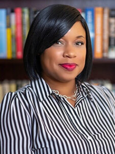 Headshot of Dr. Brandy McNeil, a Black woman with short, mid-length hair wearing a striped, black and white blouse. There is a backdrop of colourful book shelves behind her.