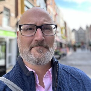 A headshot of David Lankes with a gray beard, wearing black-rimmed glasses, wearing a blue cardigan over a pink shirt. He is standing in front of a row of shops. David is a white, middle-aged male.