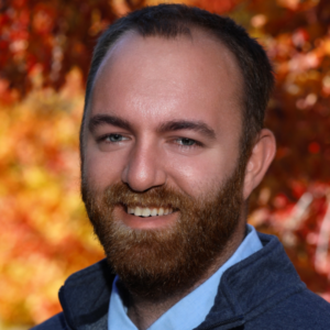 A headshot of Reed C. Hepler with a background of orange fall leaves. Reed is a white, middle-aged man with short hair and tidy beard. He is wearing a grey jacket over a collared shirt.