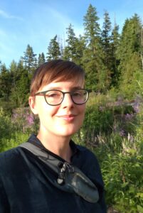 A head shot of Anne Pasek who is Caucasian, has short brown hair, and is wearing glasses. She is located in front of a forest.