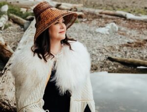 Carolyn Roberts, an Indigenous woman wears a cedar woven hat and white, fur-lined jacket. She has mid-length dark hair and stands in front of rocks and a pool of water.