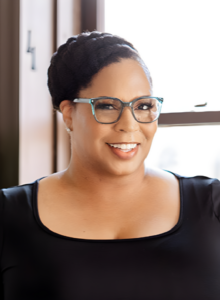 A headshot of Kaetrena Davis Kendrick who is a Black woman wearing glasses, smiling, with short hair pulled back. She wears a black top and is in front of a window.