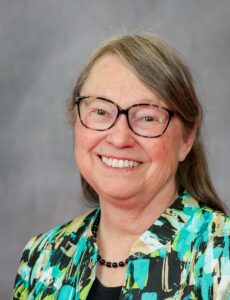 A head shot of Marydee Ojala smiling in front of a gray background. Marydee is an older white woman with long brown hair in a side part, she wears glasses and a blouse that has splashes of colour in teal, light green, and black.