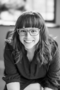 A black and white headshot photo of Jenne. She is smiling, wearing glasses and a dark blouse. Jenne has a white complexion, her hair is mid-length with bangs.