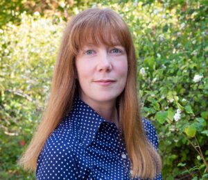 A headshot of Krista Boehnert, who is a middle-aged white woman with long, straight auburn hair and bangs. She wears a blue shirt with white polka dots, and is standing in front of a green landscape.