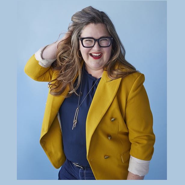 Avery Swartz is standing in front of a light blue background. She is a white, middle-aged woman with shoulder-length hair in blond and brown tones. She is smiling, wearing glasses, and has a mustard coloured sports jacket over a blue shirt and blue jeans. She wears a contemporary, long gold necklace and hoop earrings.