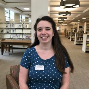 A headshot of Hannah Cyrus seated in a library. Hannah is a young white woman with shoulder length brown hair. She is smiling and wearing a blue blouse with white polka dots